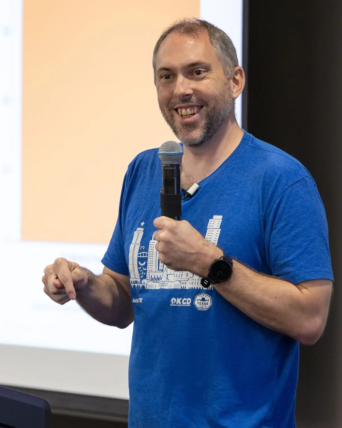 Jonathan Bryce smiles on stage at KCD Texas 2026, microphone in hand, wearing a blue KCD Texas shirt.